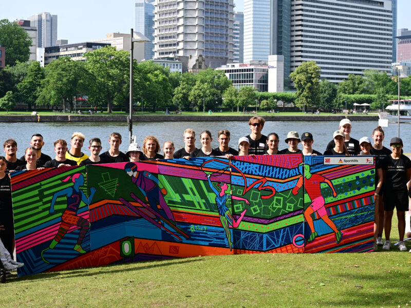Eine Gruppe von jungen Menschen und Betreuern steht vor einem farbenfroh gestalteten Fußballtor mit dem Logo der DFL Stiftung. Im Hintergrund ist die Skyline von Frankfurt zu sehen. Die Szene zeigt das NEF-Treffen der DFL Stiftung.