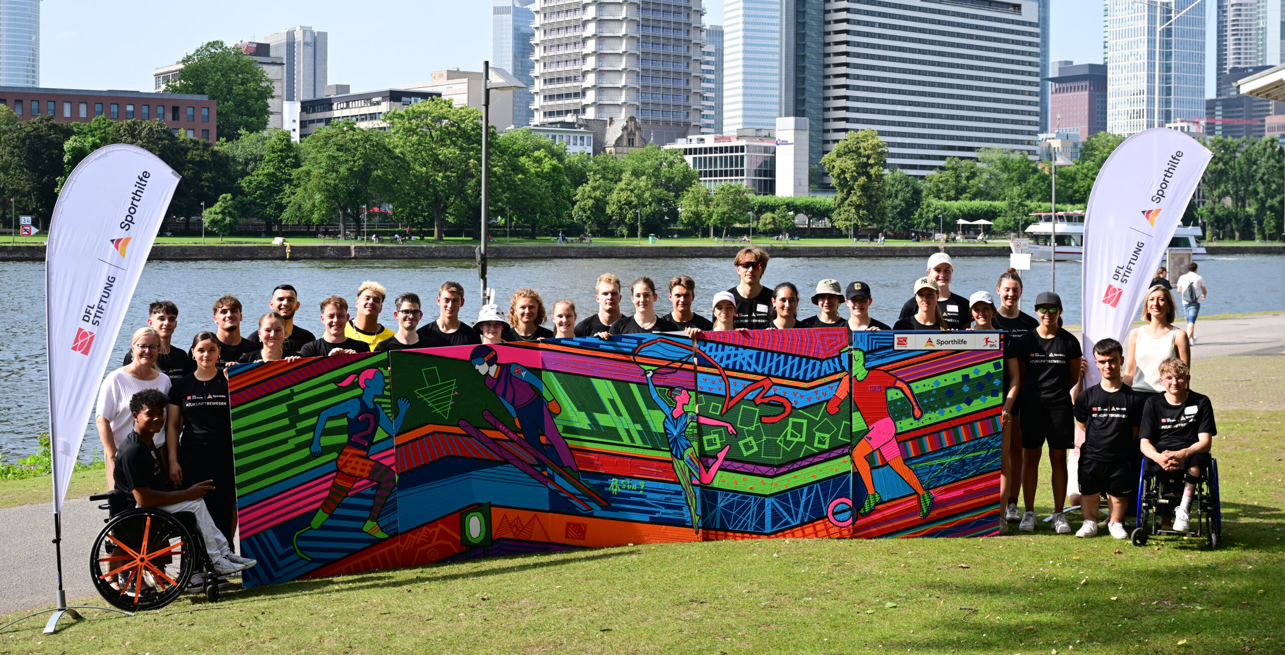 Eine Gruppe von jungen Menschen und Betreuern steht vor einem farbenfroh gestalteten Fußballtor mit dem Logo der DFL Stiftung. Im Hintergrund ist die Skyline von Frankfurt zu sehen. Die Szene zeigt das NEF-Treffen der DFL Stiftung.