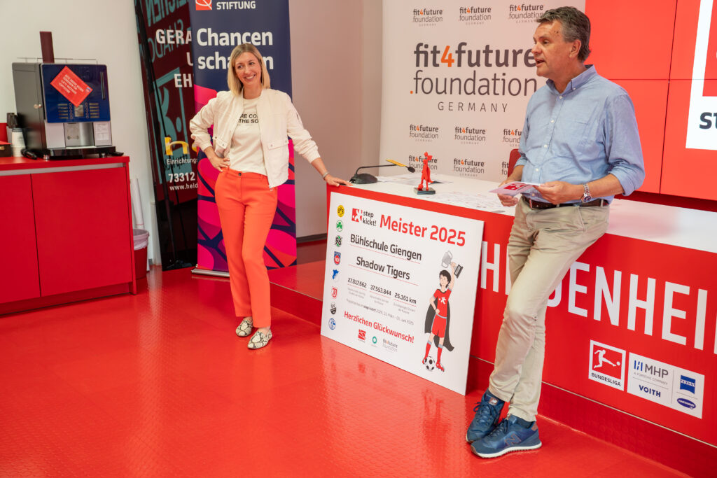 Zwei Personen überreichen einen symbolischen Scheck mit der Aufschrift "Meister 2025" an die DFL Stiftung. Im Hintergrund ist ein Banner mit dem Logo der DFL Stiftung und der Aufschrift "fit4future foundation GERMANY" zu sehen.