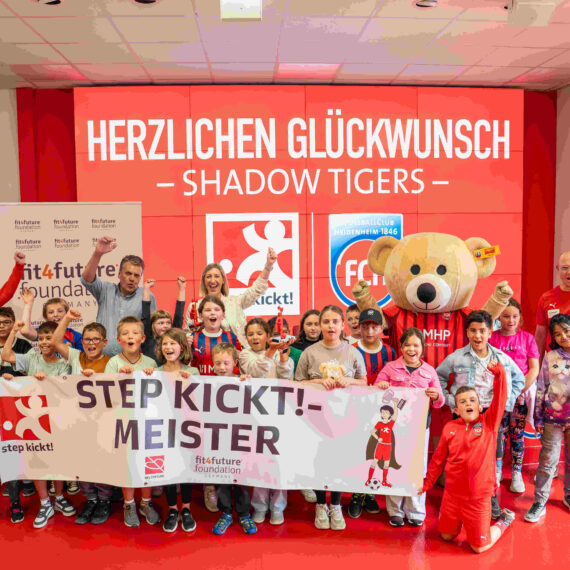 Eine Gruppe von Kindern und Jugendlichen steht vor einem roten Banner mit der Aufschrift "STEP KICKT! - MEISTER". Sie halten Banner und tragen rote Trainingsanzüge. Im Hintergrund steht ein großer Teddybär und ein Banner mit dem Logo der fitfuture foundation.