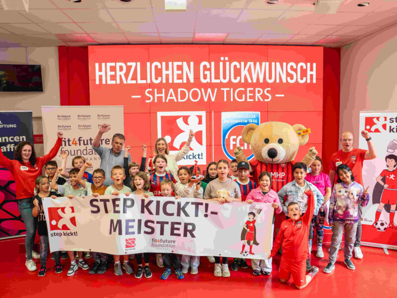 Eine Gruppe von Kindern und Jugendlichen steht vor einem roten Banner mit der Aufschrift "STEP KICKT! - MEISTER". Sie halten Banner und tragen rote Trainingsanzüge. Im Hintergrund steht ein großer Teddybär und ein Banner mit dem Logo der fitfuture foundation.