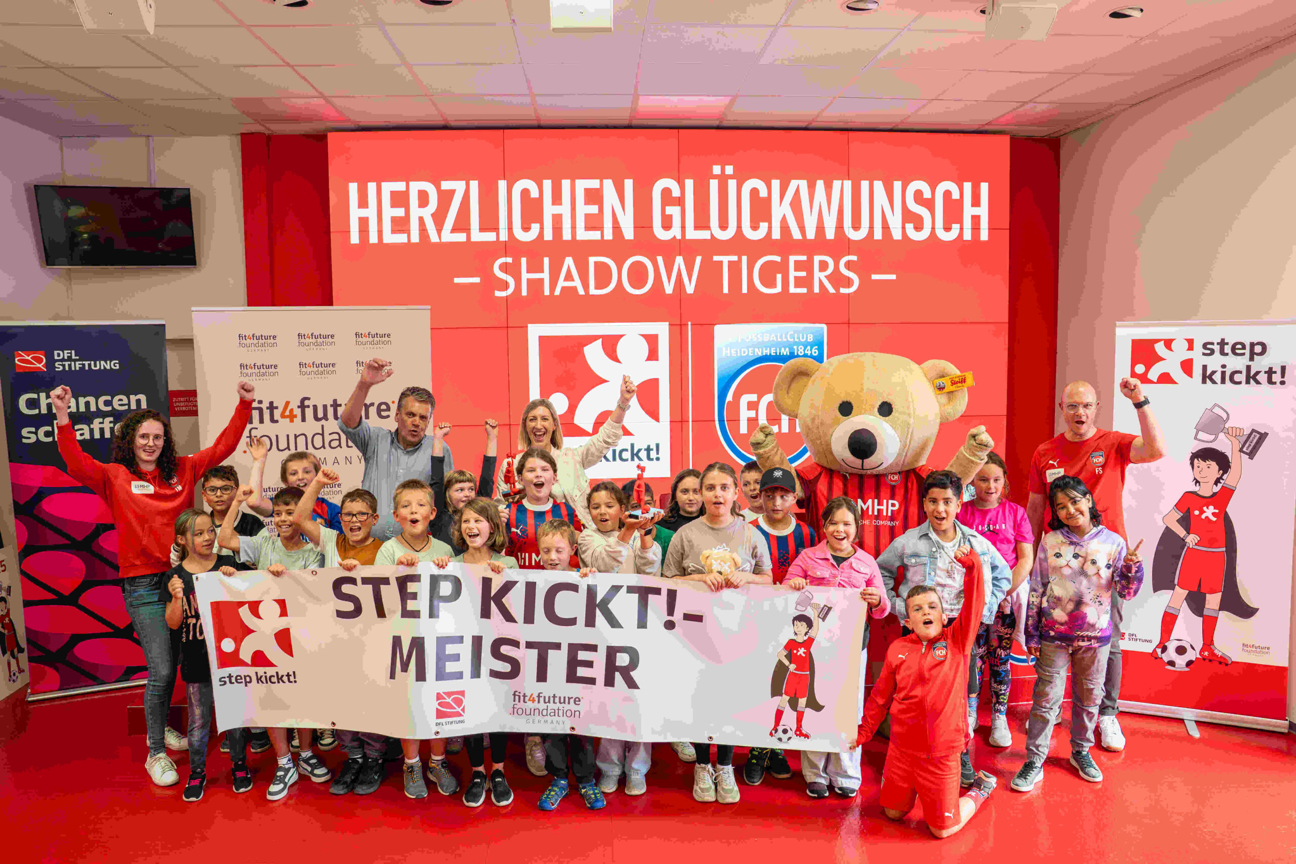 Eine Gruppe von Kindern und Jugendlichen steht vor einem roten Banner mit der Aufschrift "STEP KICKT! - MEISTER". Sie halten Banner und tragen rote Trainingsanzüge. Im Hintergrund steht ein großer Teddybär und ein Banner mit dem Logo der fitfuture foundation.