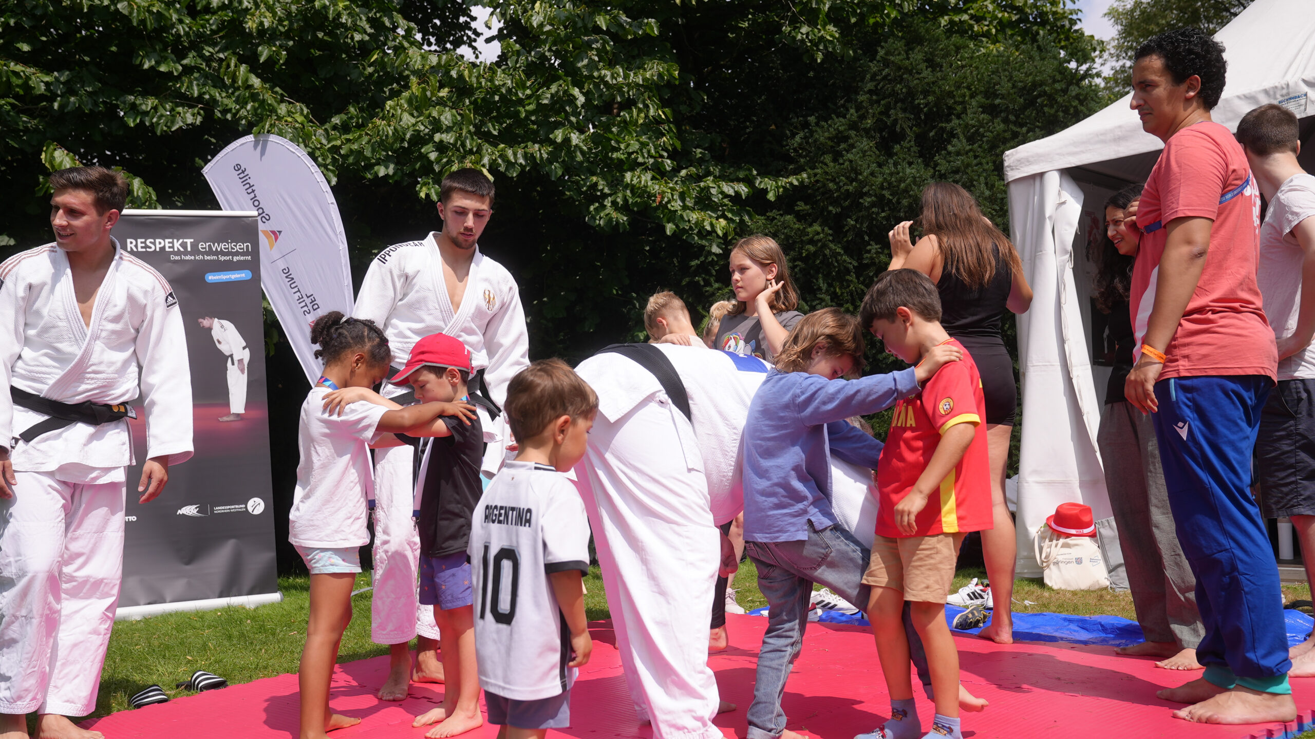 Eine Gruppe von Kindern und Jugendlichen übt gemeinsam Kampfsporttechniken. Ein Jugendlicher im weißen Judogi führt die Übungen vor, während ein Kind im argentinischen Trikot und ein anderes in roter Kleidung die Bewegungen nachahmen. Im Hintergrund sind weitere Kinder und Jugendliche zu sehen.