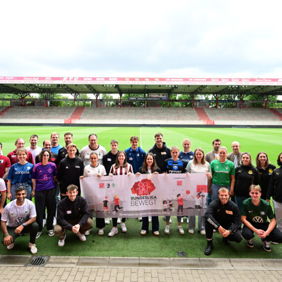 Eine Gruppe von rund 30 Personen steht auf dem Rasen eines Fußballstadions zusammen und lächelt in die Kamera. Sie tragen Kleidung mit verschiedenen Vereinslogos und der Aufschrift "Bundesliga bewegt". Im Hintergrund sind die leeren Zuschauerränge des Stadions zu sehen.