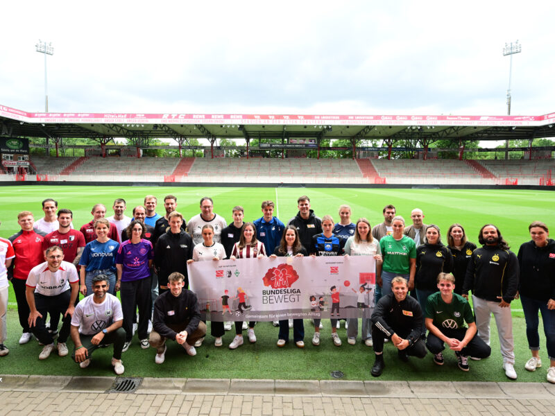 Eine Gruppe von rund 30 Personen steht auf dem Rasen eines Fußballstadions zusammen und lächelt in die Kamera. Sie tragen Kleidung mit verschiedenen Vereinslogos und der Aufschrift "Bundesliga bewegt". Im Hintergrund sind die leeren Zuschauerränge des Stadions zu sehen.
