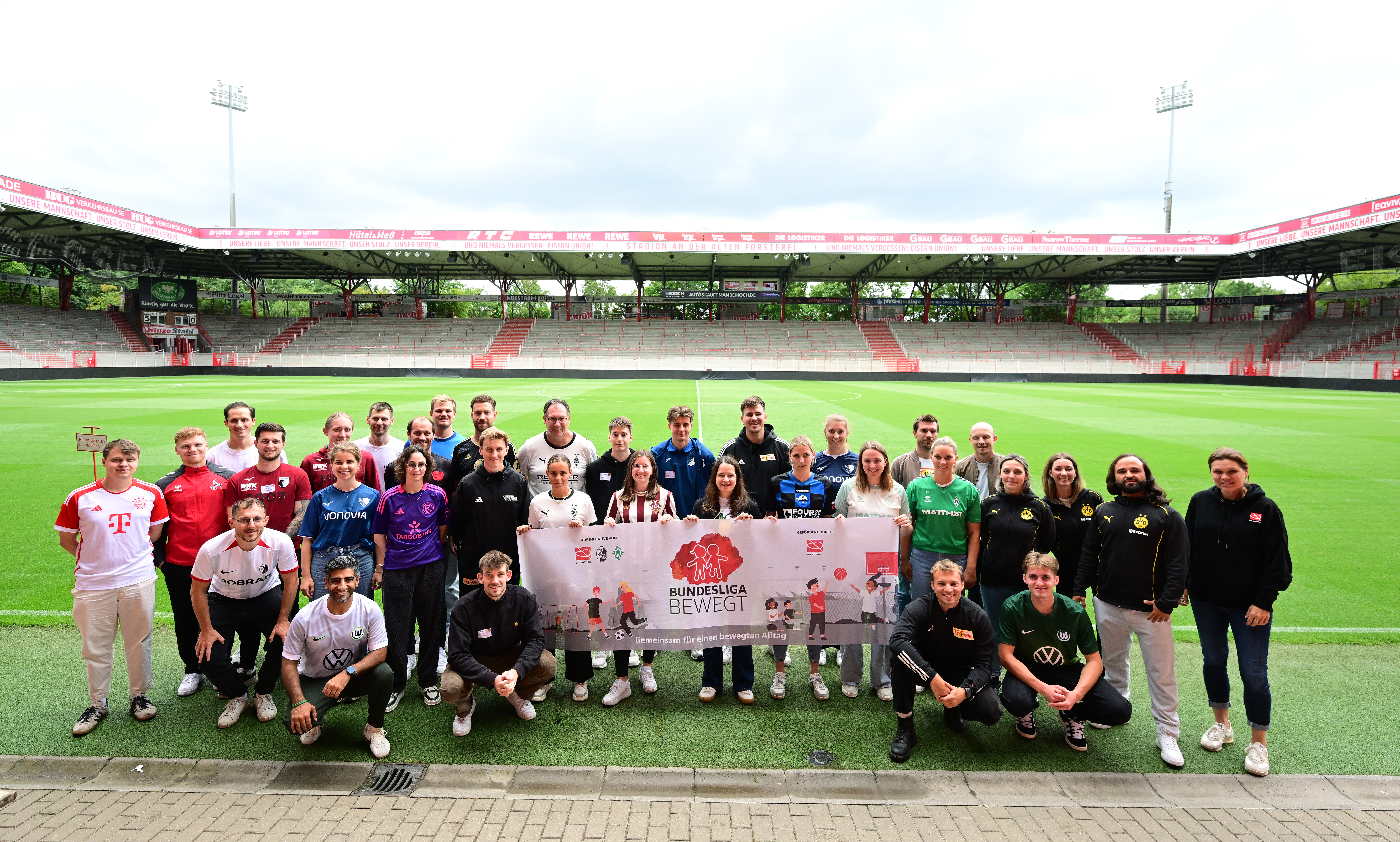 Eine Gruppe von rund 30 Personen steht auf dem Rasen eines Fußballstadions zusammen und lächelt in die Kamera. Sie tragen Kleidung mit verschiedenen Vereinslogos und der Aufschrift "Bundesliga bewegt". Im Hintergrund sind die leeren Zuschauerränge des Stadions zu sehen.