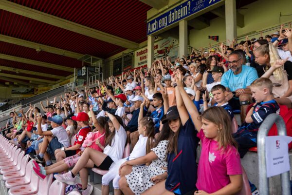 Jubelnde Kinder und Jugendliche sitzen auf den Zuschauertribünen eines Fußballstadions und recken die Arme hoch. Im Hintergrund sind weitere Zuschauer und Werbebanner zu sehen.