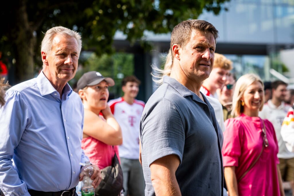 Zwei Männer in Hemden stehen mit dem Rücken zum Betrachter und unterhalten sich inmitten einer Menschenmenge bei einer Veranstaltung im Freien. Im Hintergrund sind weitere Personen und Teile der MHPArena in Stuttgart zu erkennen.