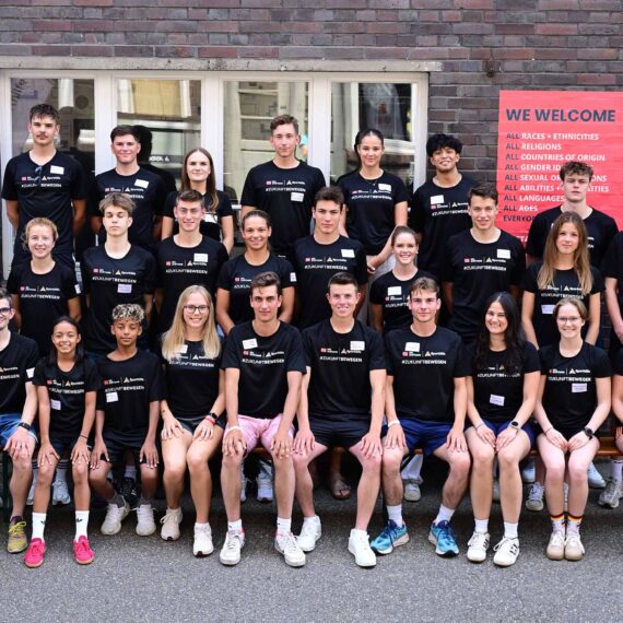 Eine Gruppe junger Fußballspieler und -spielerinnen steht für ein Gruppenfoto vor einem Gebäude. Viele tragen schwarze Trainingsanzüge mit dem Logo der DFL Stiftung und der Deutschen Sporthilfe. Im Hintergrund sind Banner mit Aufschriften wie "WELCOME" und "ALL RACES, ETHNICITIES" zu sehen. Ein Teilnehmer sitzt im Rollstuhl.