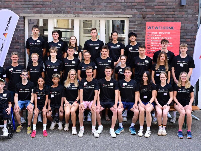 Eine Gruppe junger Fußballspieler und -spielerinnen steht für ein Gruppenfoto vor einem Gebäude. Viele tragen schwarze Trainingsanzüge mit dem Logo der DFL Stiftung und der Deutschen Sporthilfe. Im Hintergrund sind Banner mit Aufschriften wie "WELCOME" und "ALL RACES, ETHNICITIES" zu sehen. Ein Teilnehmer sitzt im Rollstuhl.