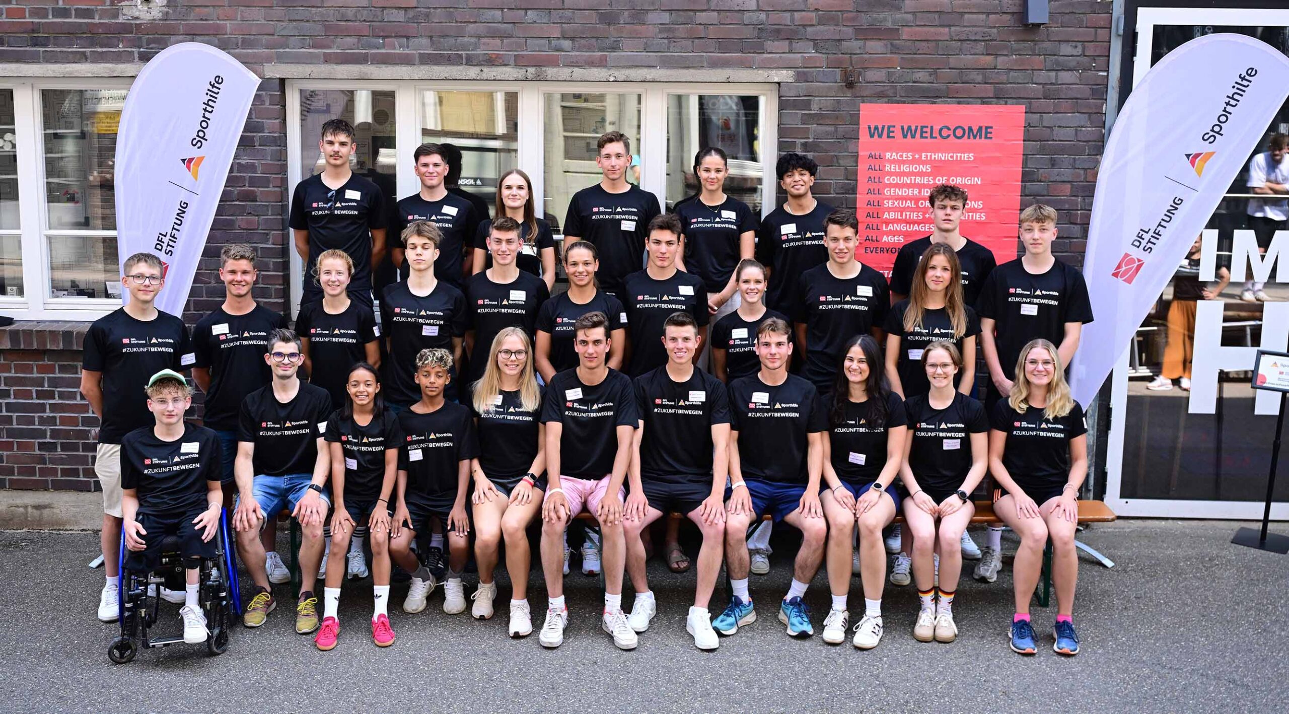 Eine Gruppe junger Fußballspieler und -spielerinnen steht für ein Gruppenfoto vor einem Gebäude. Viele tragen schwarze Trainingsanzüge mit dem Logo der DFL Stiftung und der Deutschen Sporthilfe. Im Hintergrund sind Banner mit Aufschriften wie "WELCOME" und "ALL RACES, ETHNICITIES" zu sehen. Ein Teilnehmer sitzt im Rollstuhl.