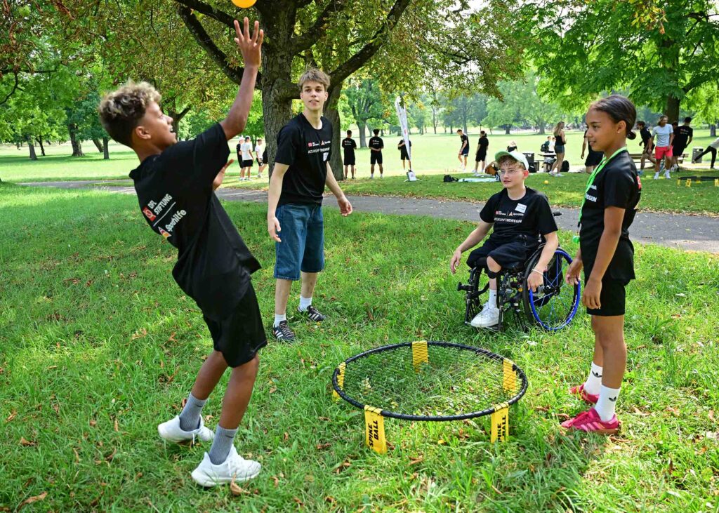 Ein Junge wirft einen Ball im Rollstuhl-Fußballtraining. Im Hintergrund stehen weitere Kinder und Jugendliche auf dem Rasen, einige nehmen am Training teil.