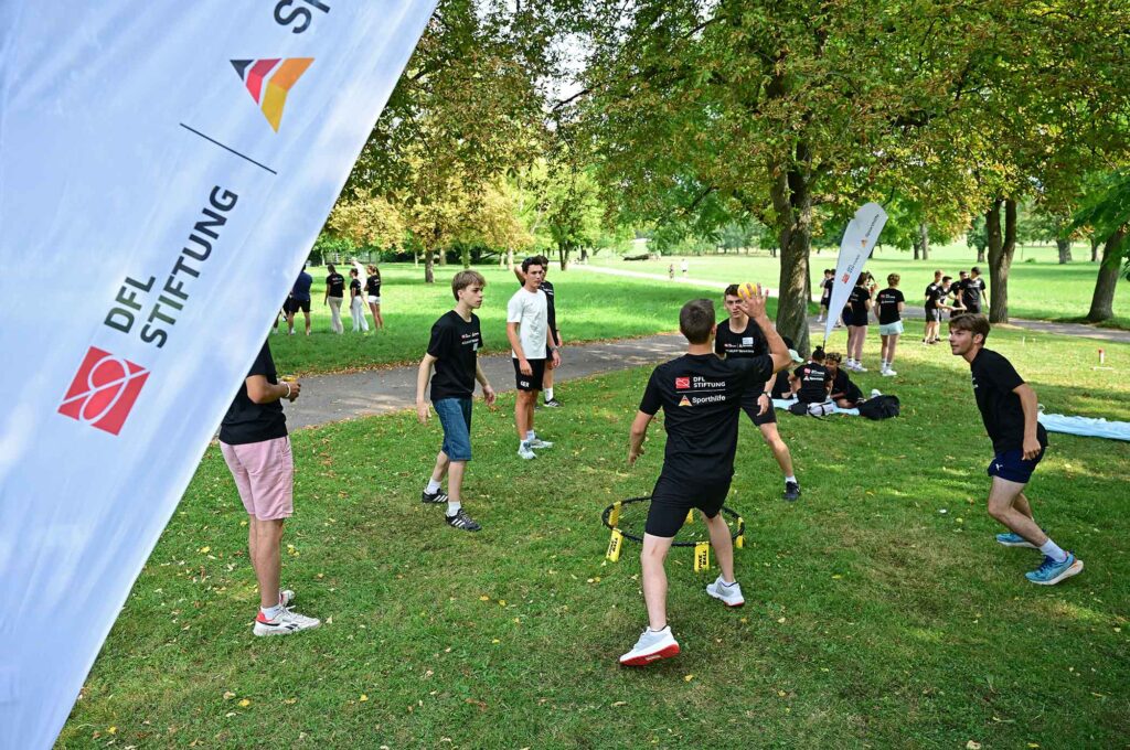 Eine Gruppe von Jugendlichen und jungen Erwachsenen nimmt an einer Fußball-Koordinationseinheit im Freien teil. Sie bewegen sich auf einem grünen Rasen, während ein Banner mit dem Logo der DFL Stiftung im Vordergrund zu sehen ist.
