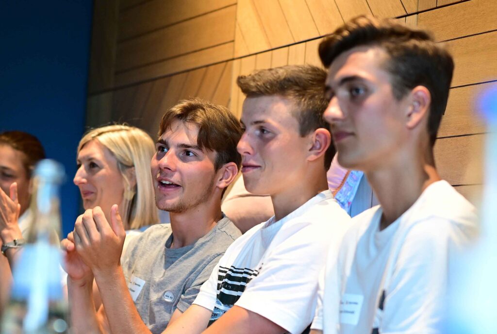 Eine Gruppe junger Fußballspieler sitzt an einem Tisch und applaudiert. Im Hintergrund ist eine Holzverkleidung zu sehen. Das Bild zeigt eine Szene vom Nachwuchselite-Treffen der DFL Stiftung und der Deutschen Sporthilfe in Stuttgart.