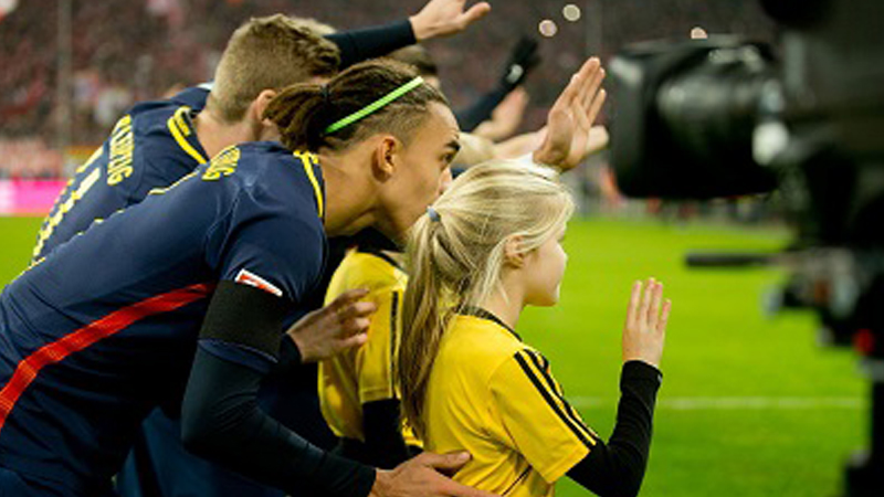 Der Profifußballer Yussuf Poulsen von RB Leipzig steht hinter einem Einlaufkind, welches den Fans und Kameras zuwinkt.