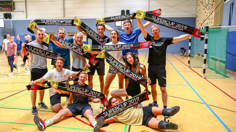 Elf Nachwuchselite-Athletinnen und -Athleten stehen für ein Gruppenfoto in einer Turnhalle zusammen. Sie halten mehrere Schals in den Händen, auf denen Supercup 2019 steht. 